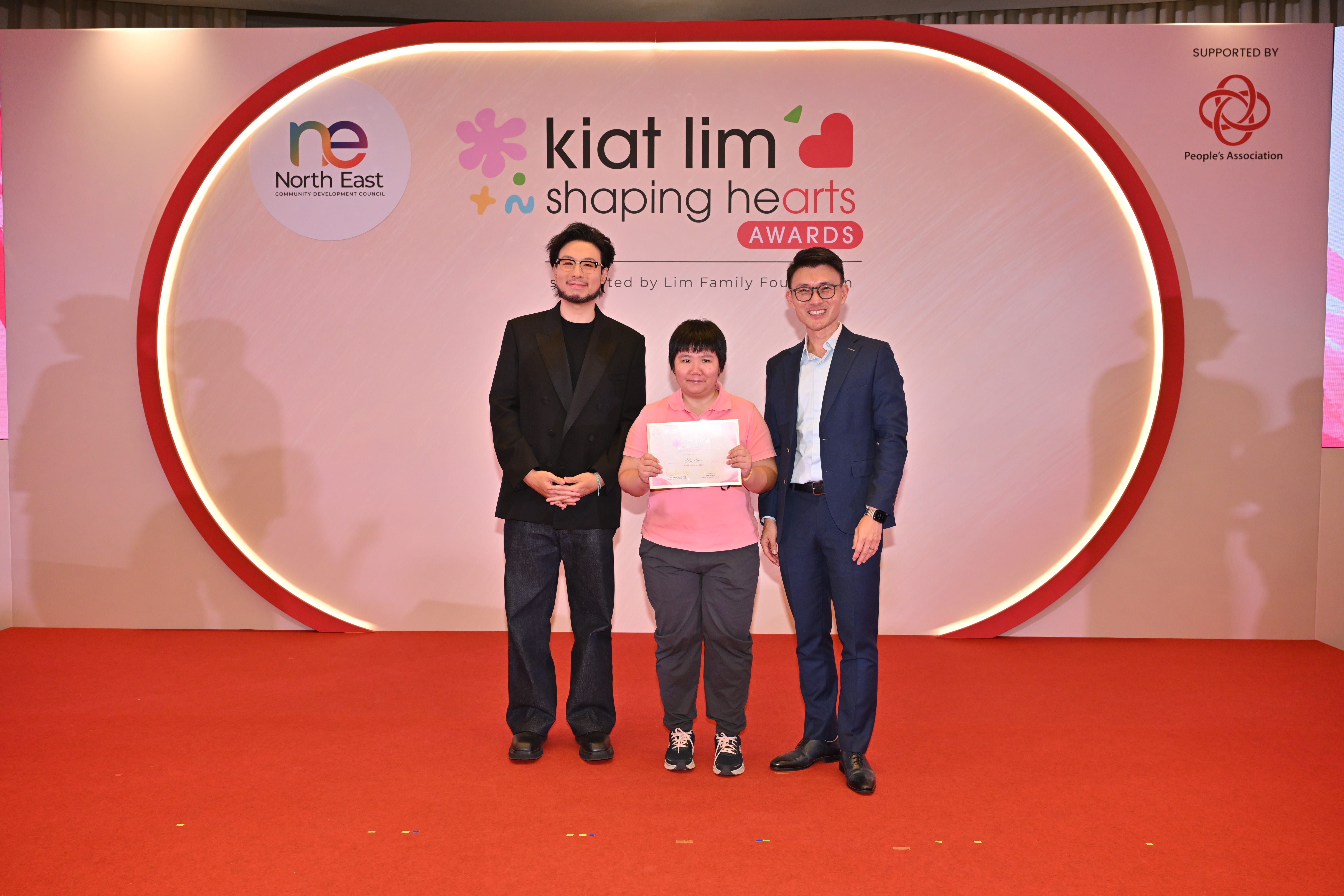 Award recipient standing on stage with the presenter, holding their certificate and posing for a group photo during the award ceremony, with a backdrop reading ‘Kiat Lim-Shaping Hearts Awards'.